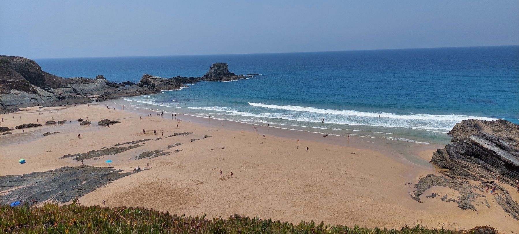 Foto do areal da praia de Zambujeira do Mar, a maré está baixa, o areal liso e enorme. À direita e à esquerda há rochas e o mar prolonga-se até colar com o céu no horizonte. A imagem divide-se quase na perfeição numa linha que une o canto superior esquerdo ao inferior direito, com a areia a dominar a parte inferior e o mar e o céu a superior. 